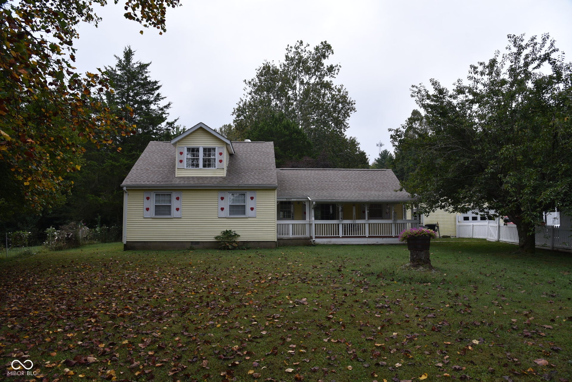 Yellow siding exterior featuring a covered front porch with white railing, dormer window, and mature surrounding trees
