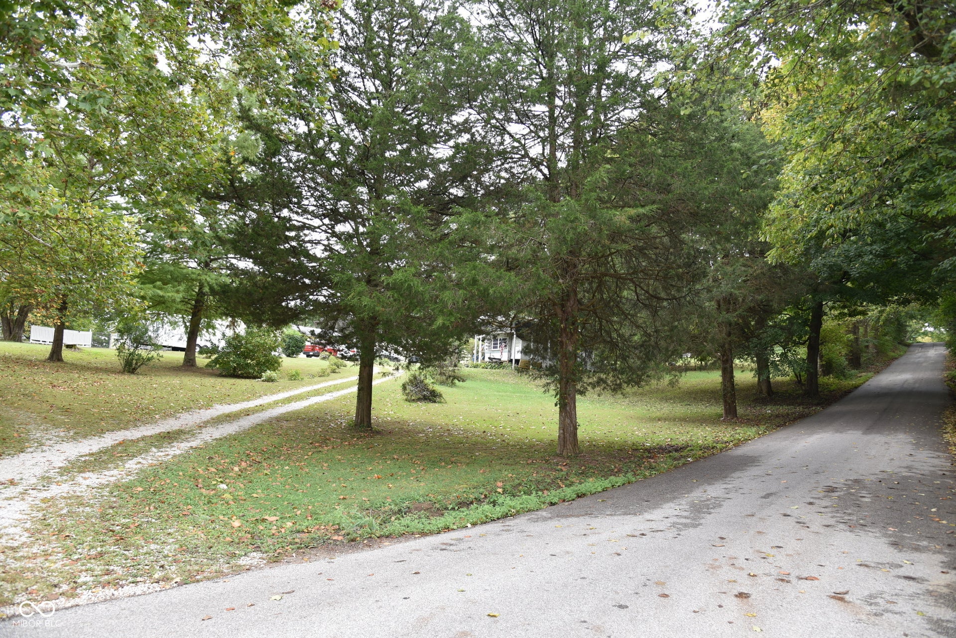 Paved road with a gravel driveway entrance, leading to a landscaped property with mature trees and a manicured lawn