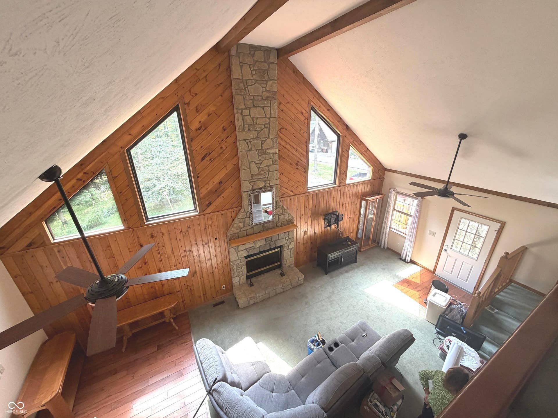 Vaulted ceiling with exposed wood beams, stone-clad fireplace, wood-finish wall paneling, and multiple transom windows