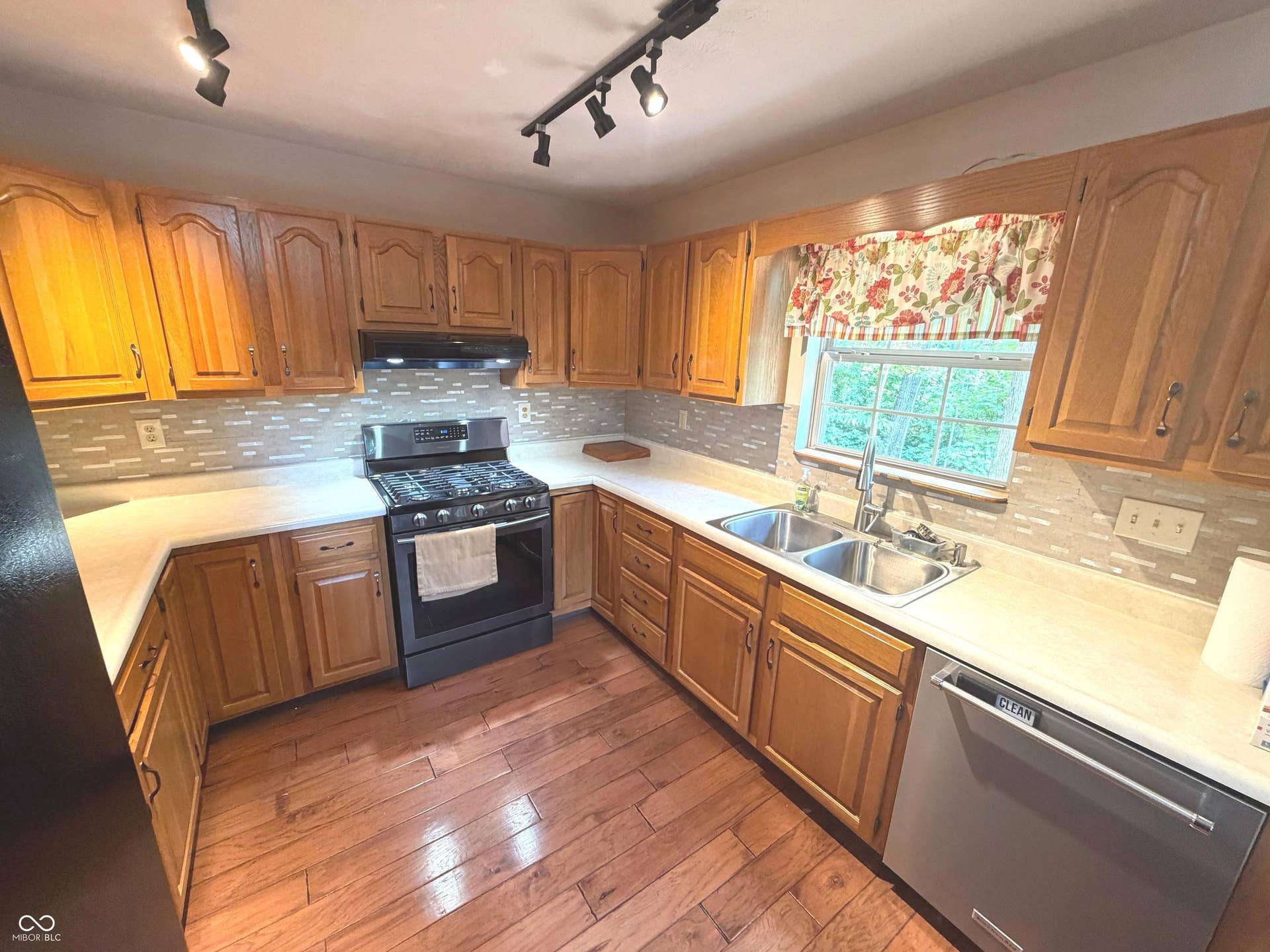 U-shaped kitchen featuring light wood cabinetry, a stainless steel gas range, a dual basin sink, wood-finish flooring, and a tile backsplash