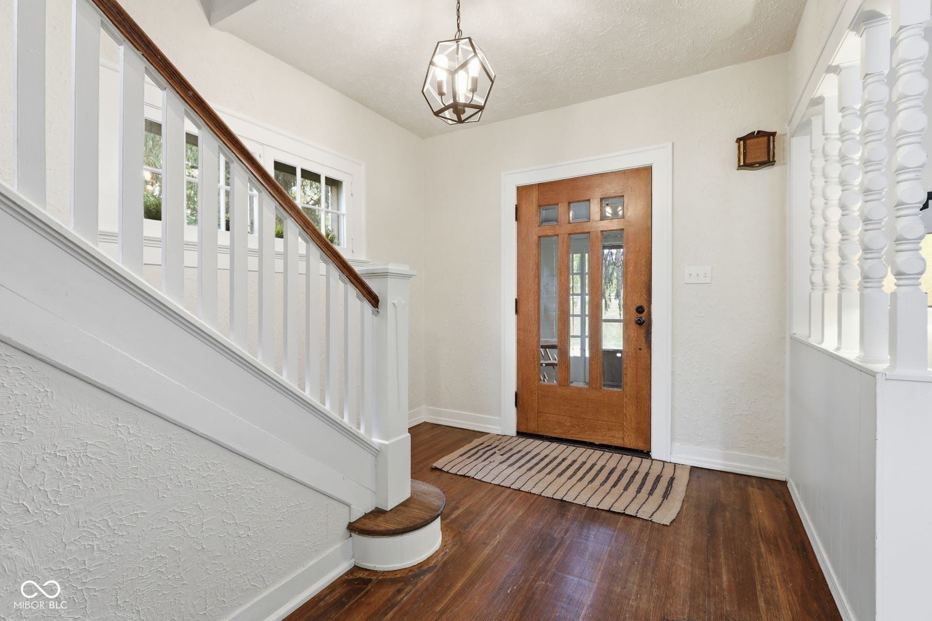 Hardwood-finish flooring, wood-paneled entry door with glass inserts, traditional white banister, overhead pendant light fixture, and textured wall finish