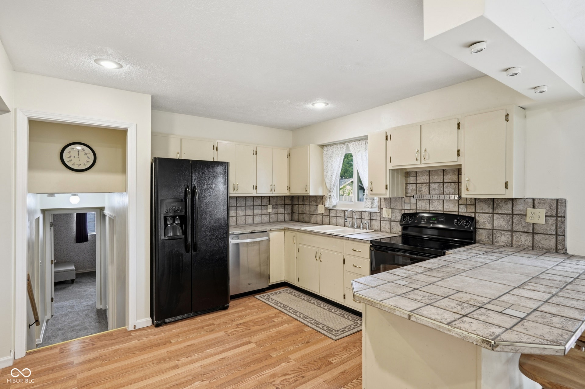 7927 Hoop Road, Indianapolis, IN 46217 Kitchen featuring wood-finish laminate flooring, white cabinetry, integrated stainless steel dishwasher, black range, and a tiled countertop with an extended breakfast bar