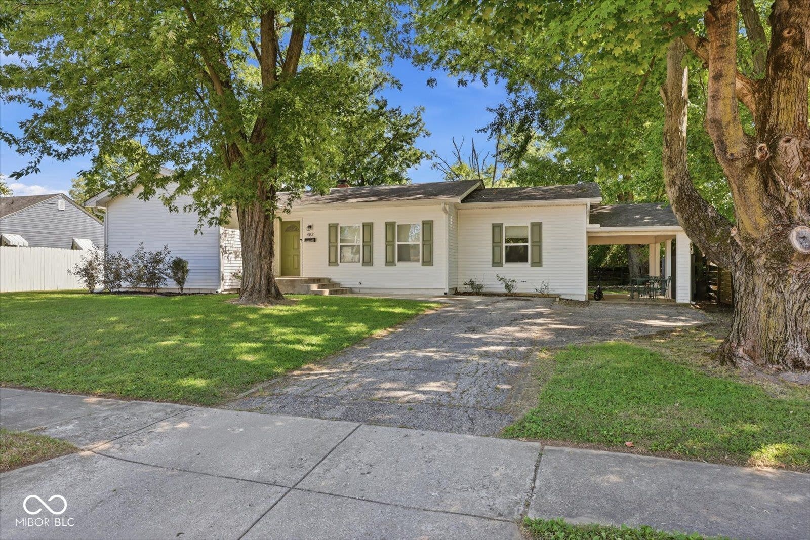 White siding exterior with green window shutters, covered carport, concrete driveway, and mature landscaping
