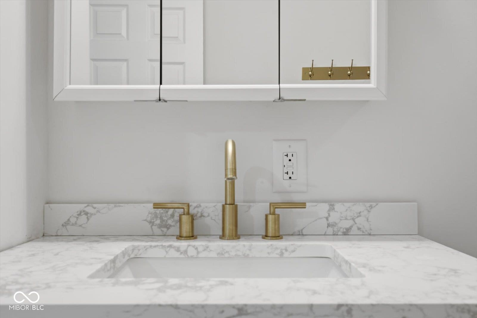 White vanity with an integrated sink, polished brass widespread faucet, and a wall-mounted mirrored medicine cabinet
