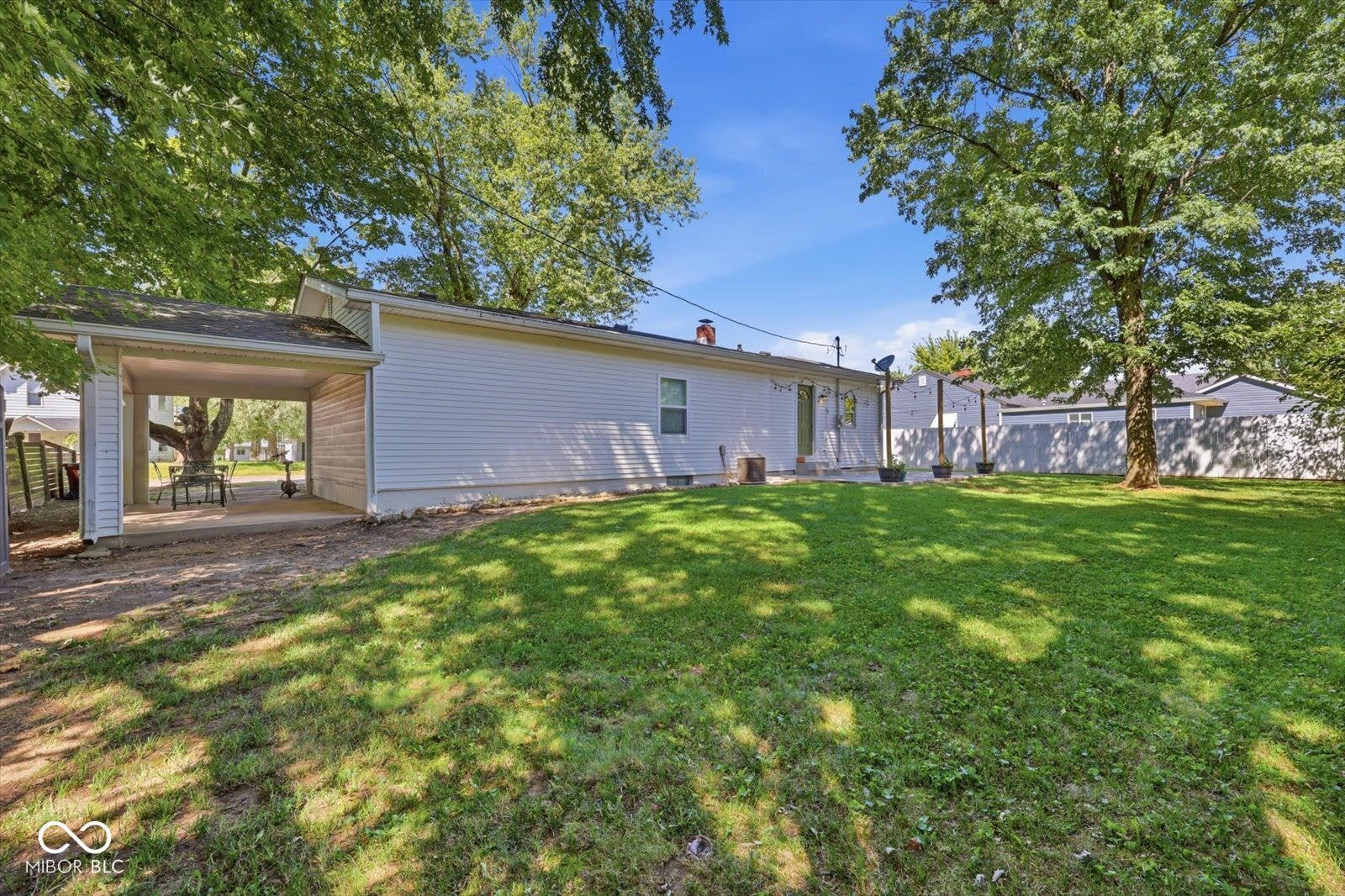 Expansive grassy yard with mature trees, a covered carport, and a white siding exterior
