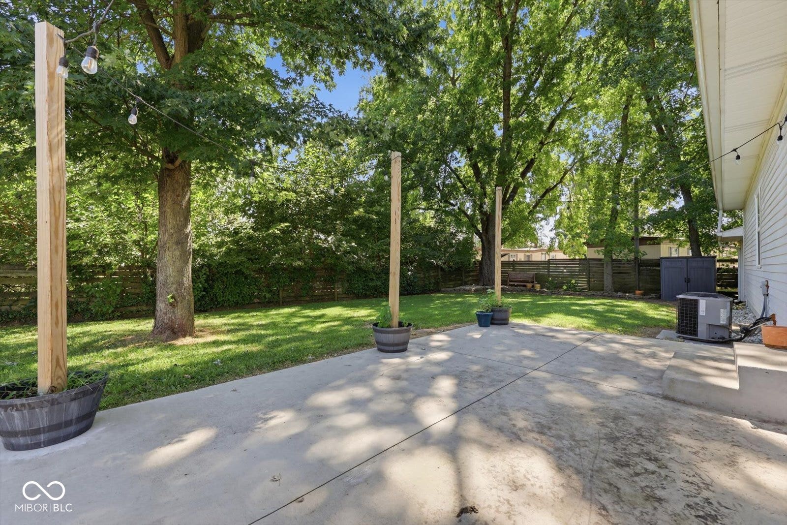 Expansive concrete patio with string lighting posts, large grassy area, and mature trees