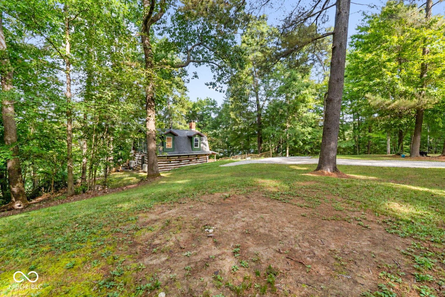 Log cabin exterior with dormer windows, surrounded by mature trees and a gravel driveway