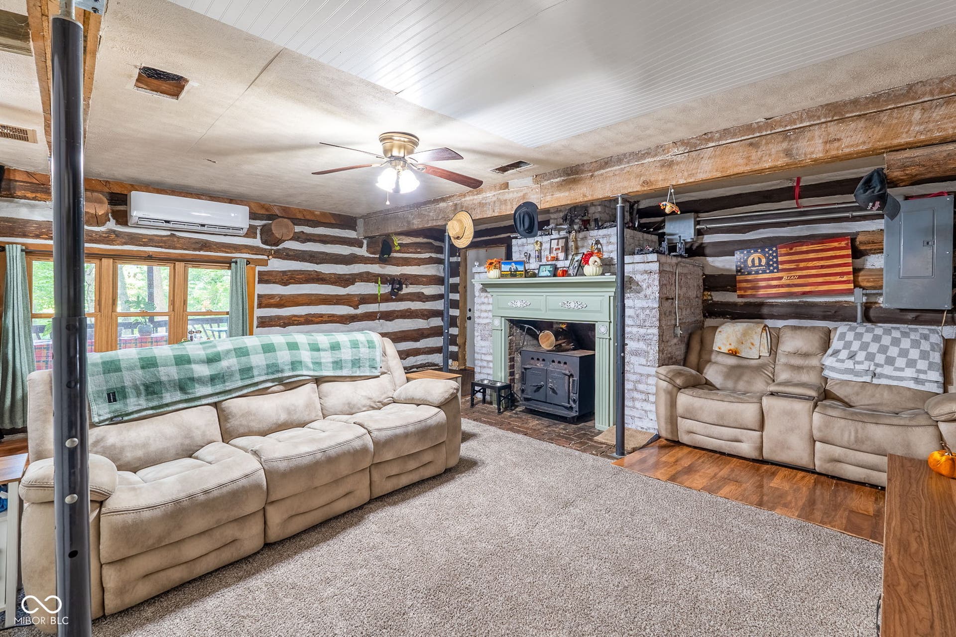 Exposed log walls, wood-finish flooring, a decorative fireplace with a wood-burning stove, and a white paneled ceiling with a ceiling fan