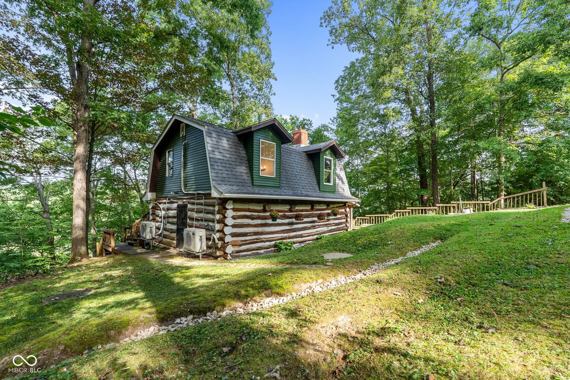 Log cabin exterior with gambrel roofline, dormer windows, and surrounding mature trees