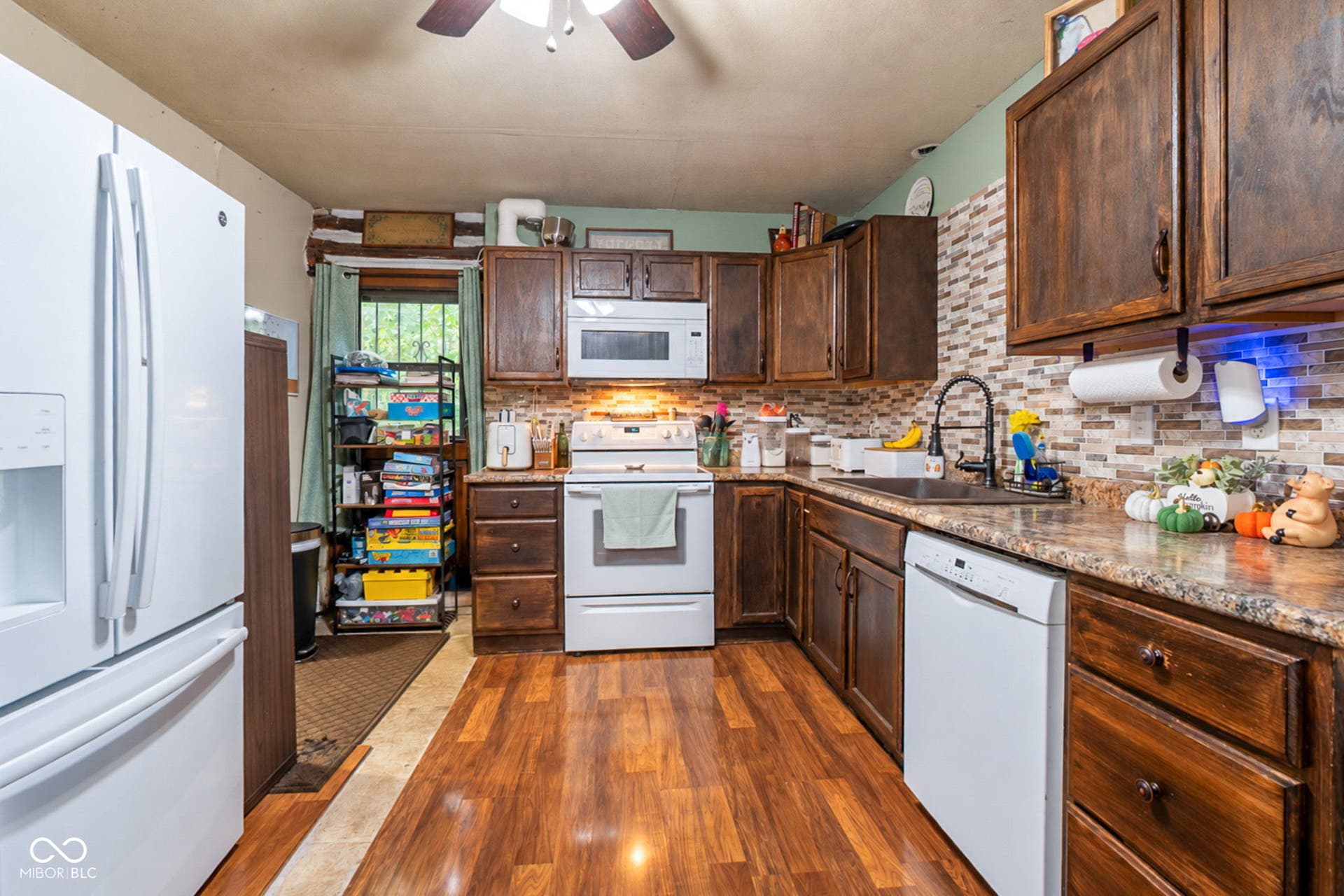Kitchen featuring wood-finish flooring, dark wood cabinetry, white appliances, a tiled backsplash, and a ceiling fan