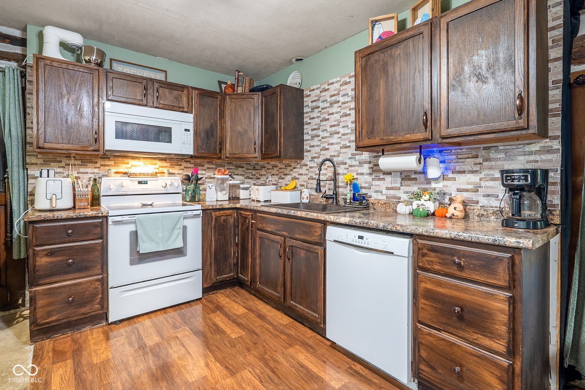 Wood-finish flooring, dark wood cabinetry, mosaic tile backsplash, countertop with integrated sink, and white appliances