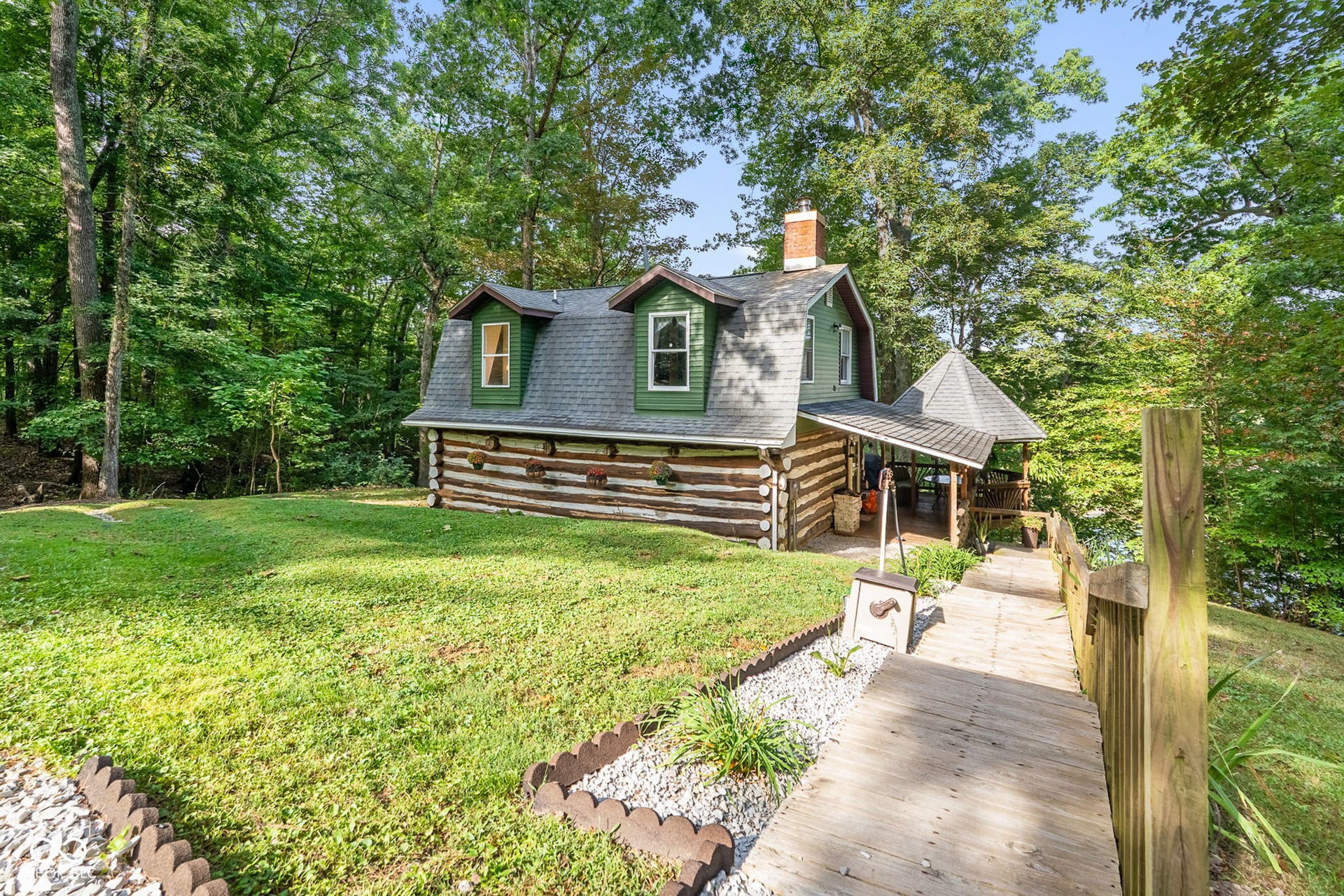Log cabin exterior with a green gambrel roof, two dormer windows, a brick chimney, an attached covered porch, and a wooden walkway