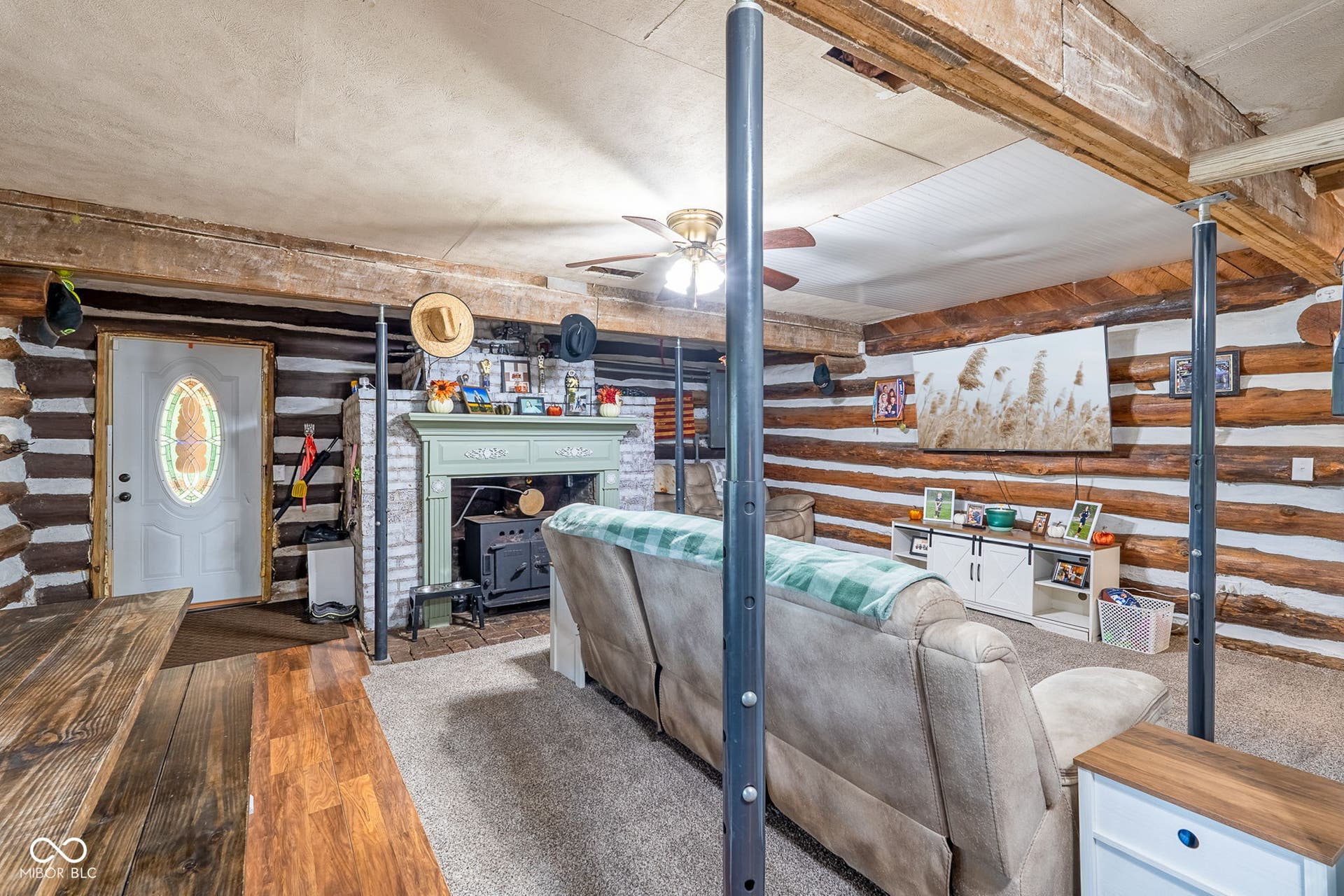 Exposed log walls, an ornate fireplace with a wood stove insert, a white entry door with an oval stained-glass insert, and a ceiling fan with integrated lighting