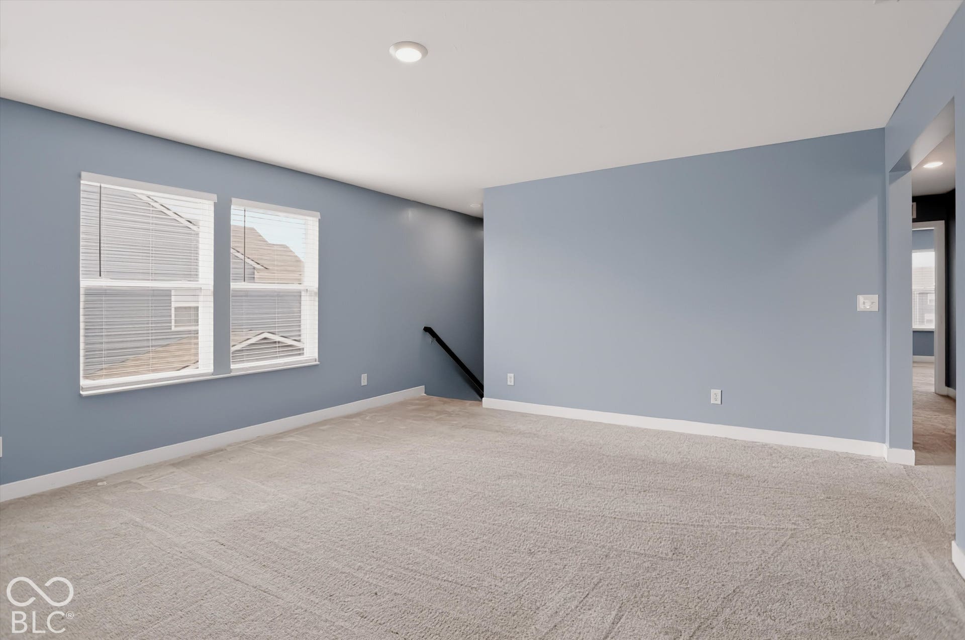 Spacious room featuring neutral carpeting, recessed ceiling lighting, dual windows with integrated blinds, and a staircase with a matte black handrail