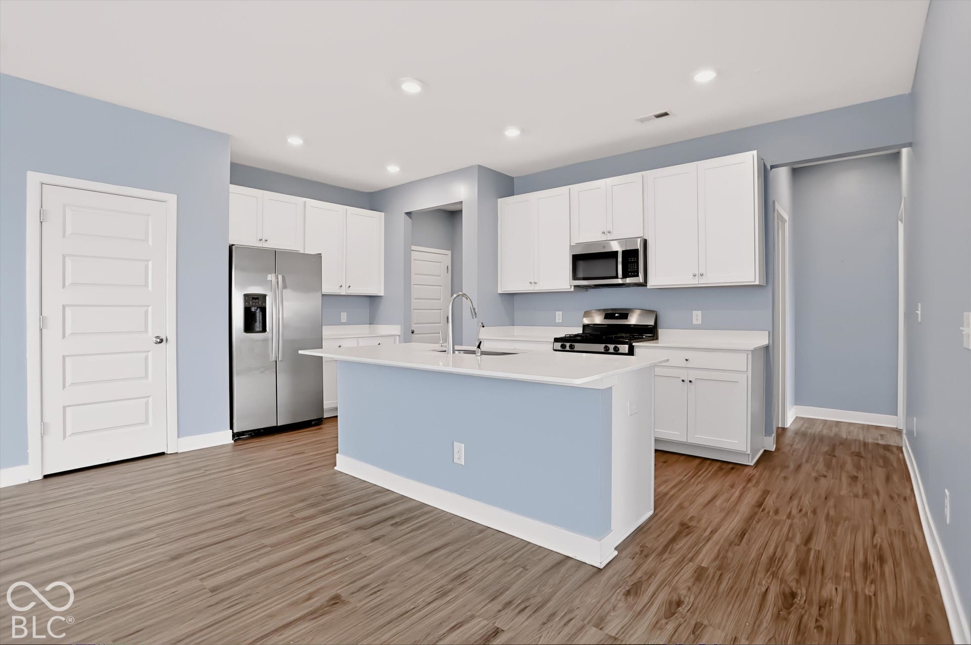 Kitchen featuring white shaker-style cabinetry, quartz countertops, stainless steel appliances, a central island with an integrated sink, and wood-finish flooring