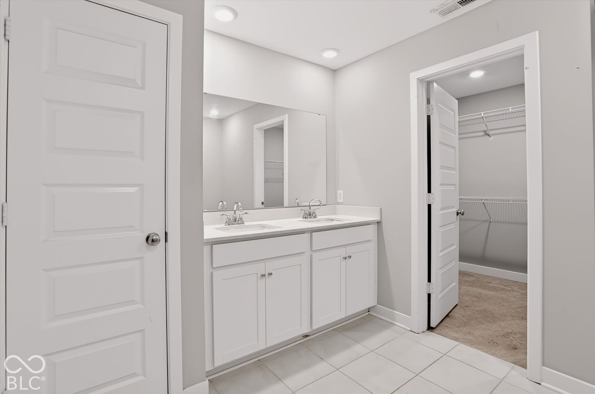 Dual-sink vanity with white cabinetry and solid-surface countertop, full-width wall mirror, recessed lighting, and walk-in closet with wire shelving