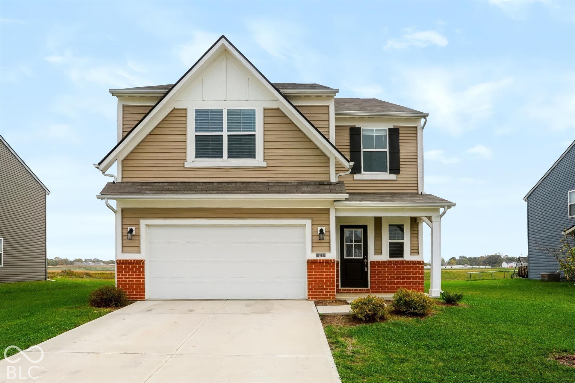 Two-story exterior featuring tan vinyl siding, a red brick veneer base, a two-car garage, and a covered front entryway with decorative shutters