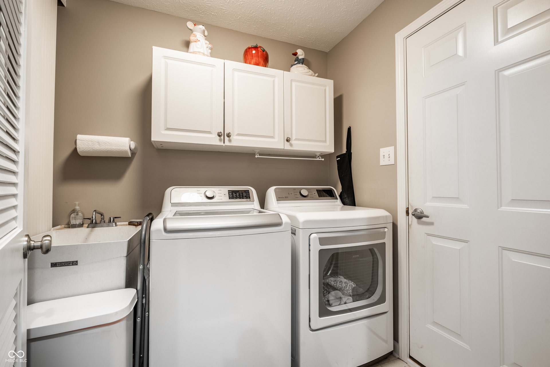 Dedicated laundry area featuring white upper cabinetry, a utility sink, and a louvered door. Leads to garage and walk thru to the kitchen.