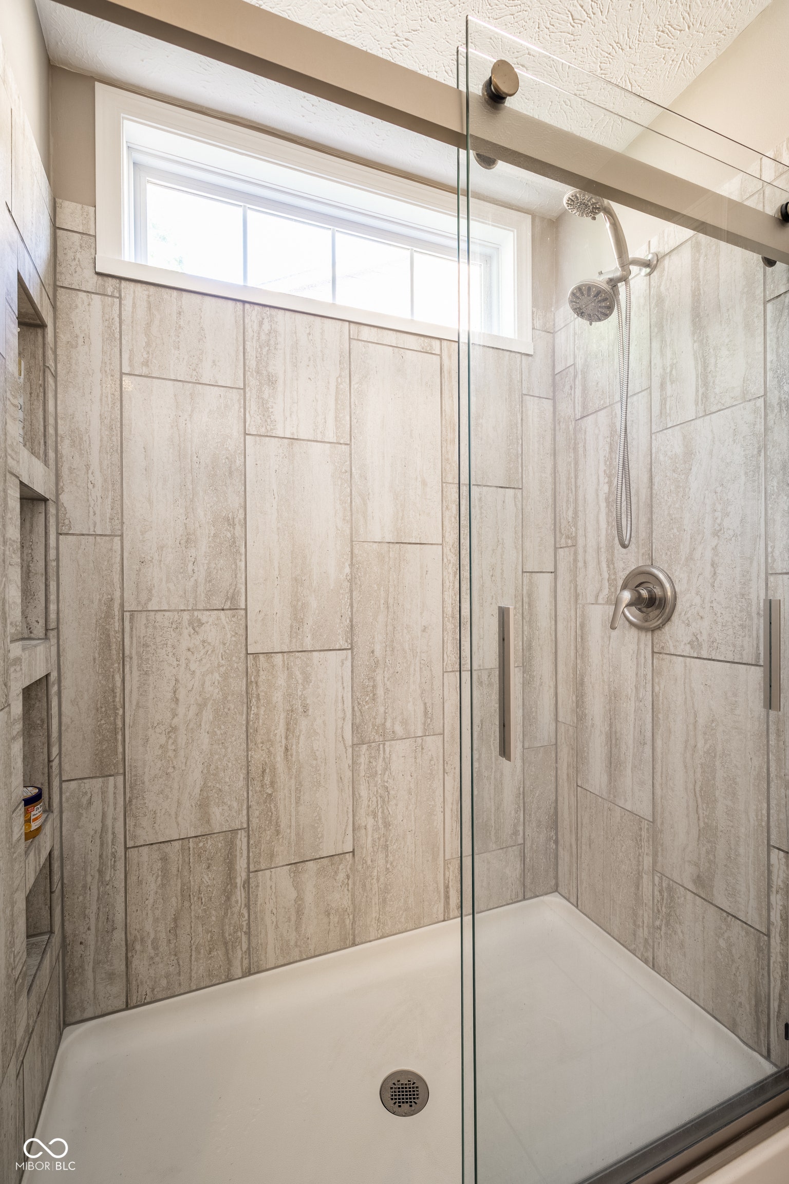 Tiled shower enclosure featuring a translucent window, glass sliding door, built-in recessed shelving, brushed nickel fixtures, and a shower wand