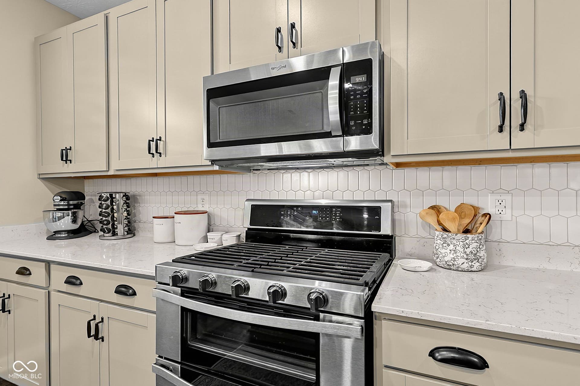 Stainless steel gas range with coordinating microwave, quartz countertops, white picket tile backsplash, and light-toned cabinetry with dark hardware
