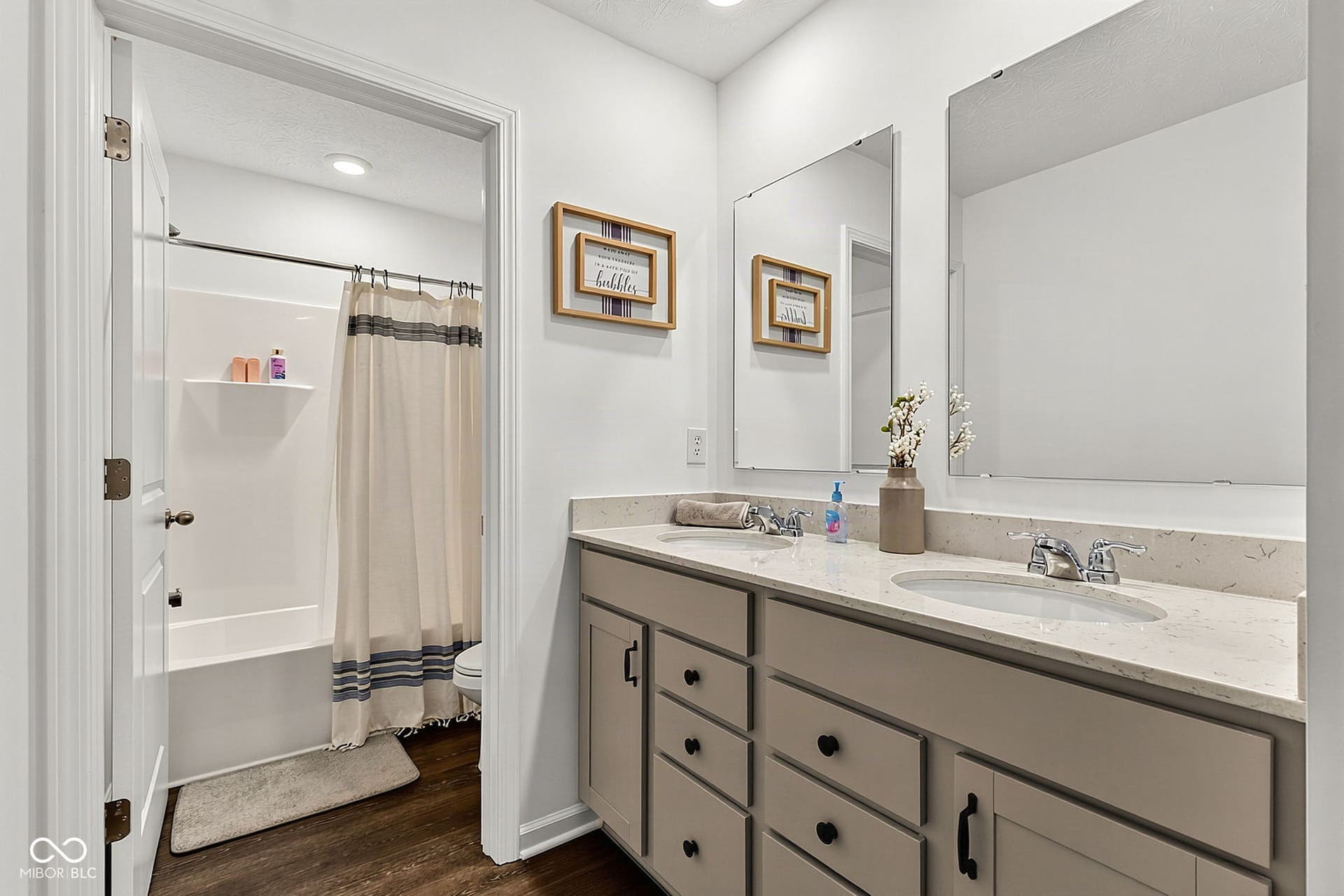 Double vanity with integrated sinks and chrome fixtures, light-toned countertop, wood-finish flooring, and a white tub with shower