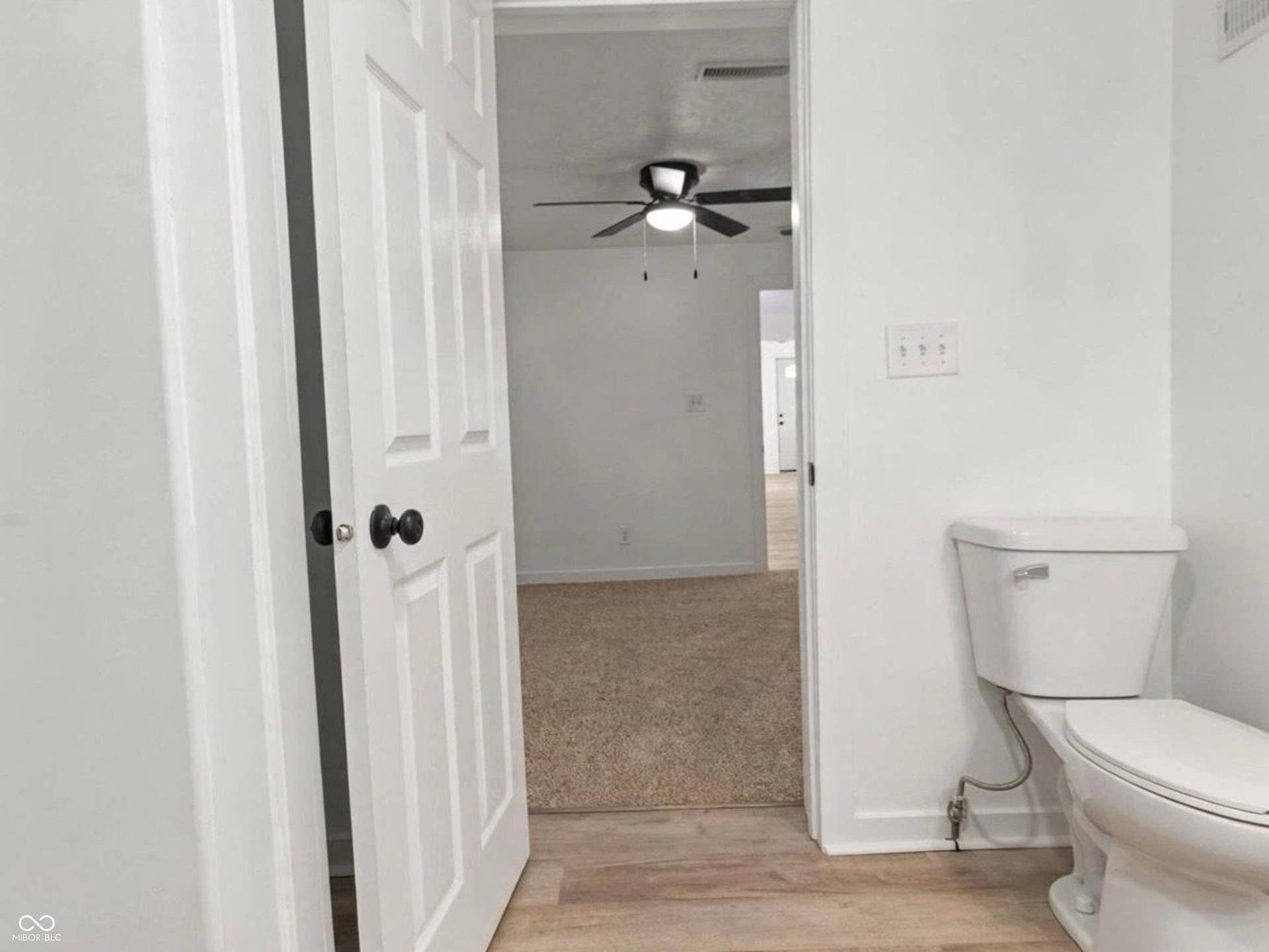 White interior door with black hardware, white toilet, light wood-finish flooring, and light-colored carpeting