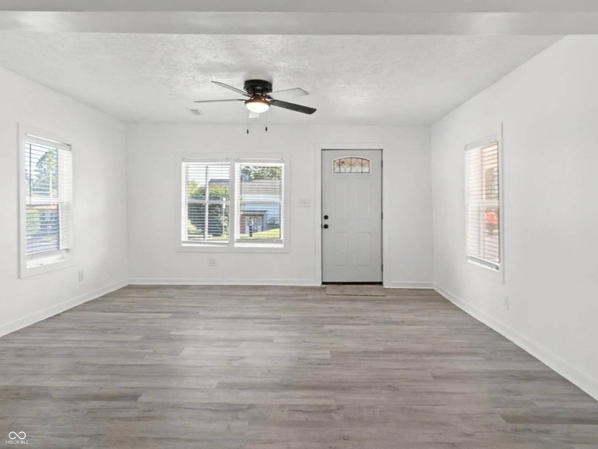 Wood-finish flooring, white walls, multiple windows with blinds, a ceiling fan, and a white entry door with decorative glass