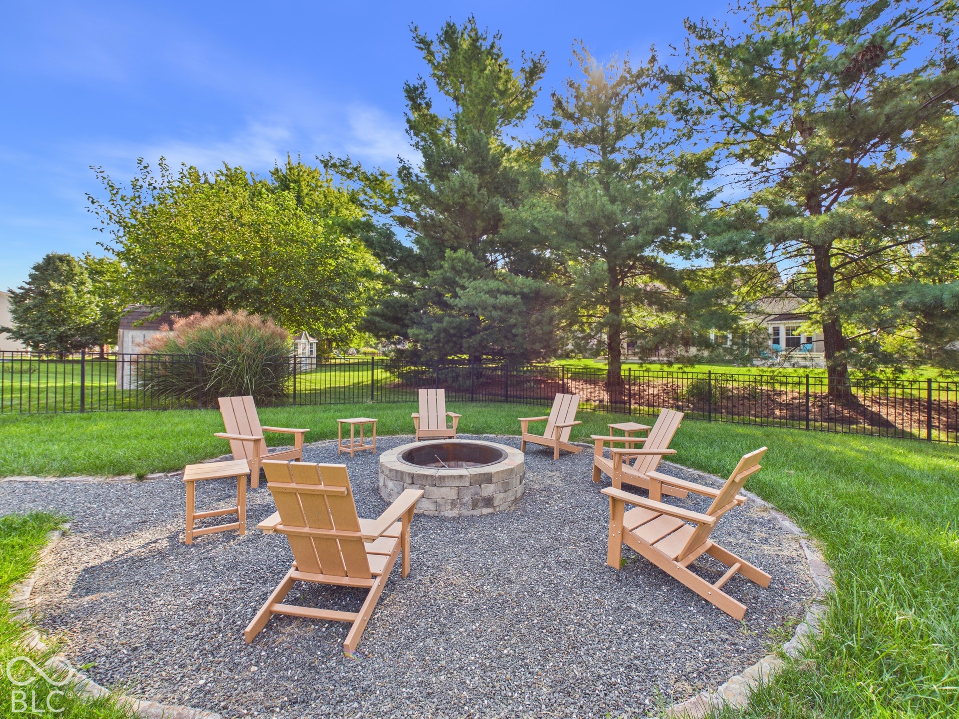 Gravel patio area featuring a stone fire pit, surrounded by mature trees, a manicured lawn, and a black fence