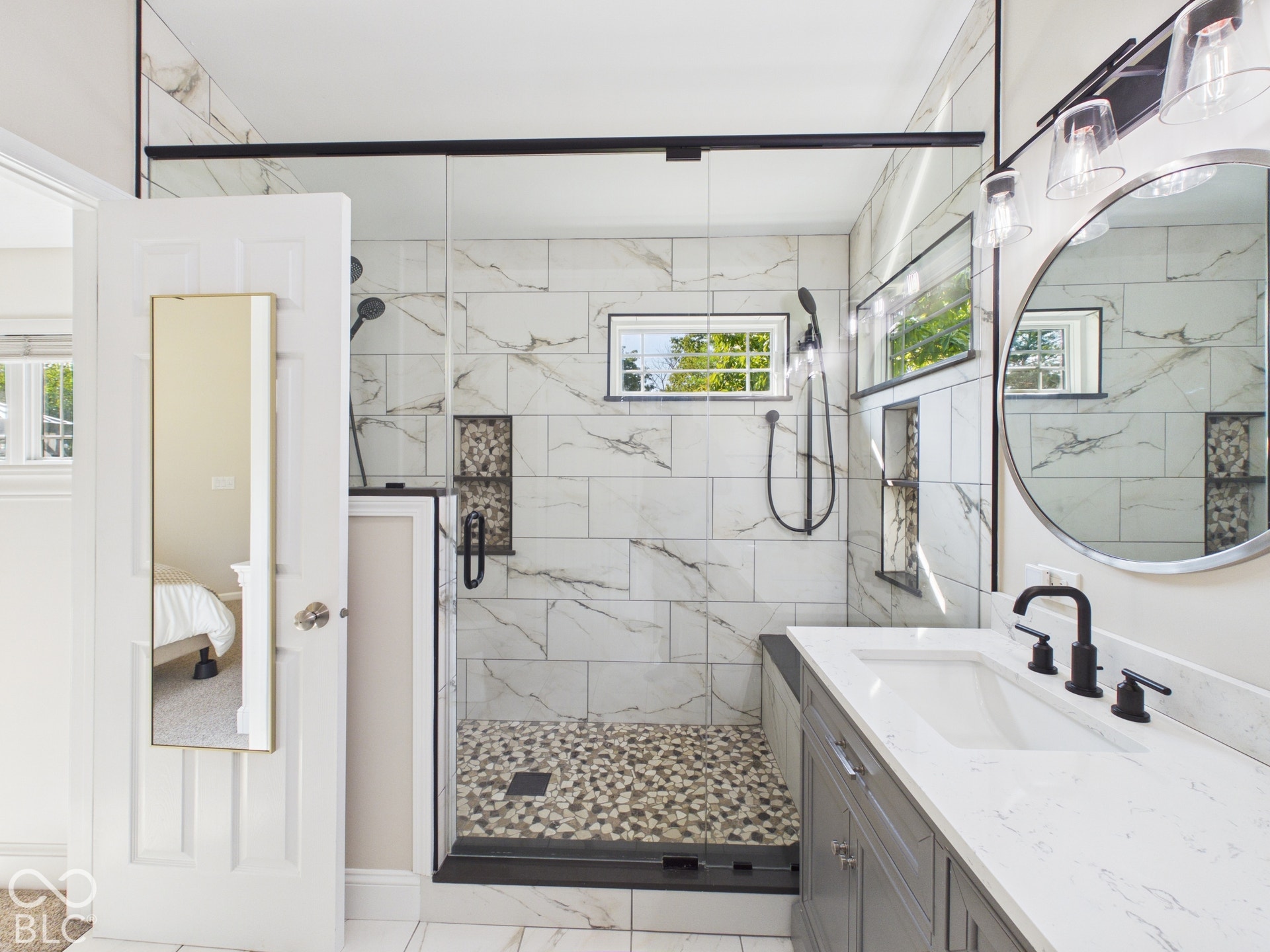 Spacious shower with glass enclosure, large format wall tiling, pebble shower floor, dual built-in shower niches, and a window