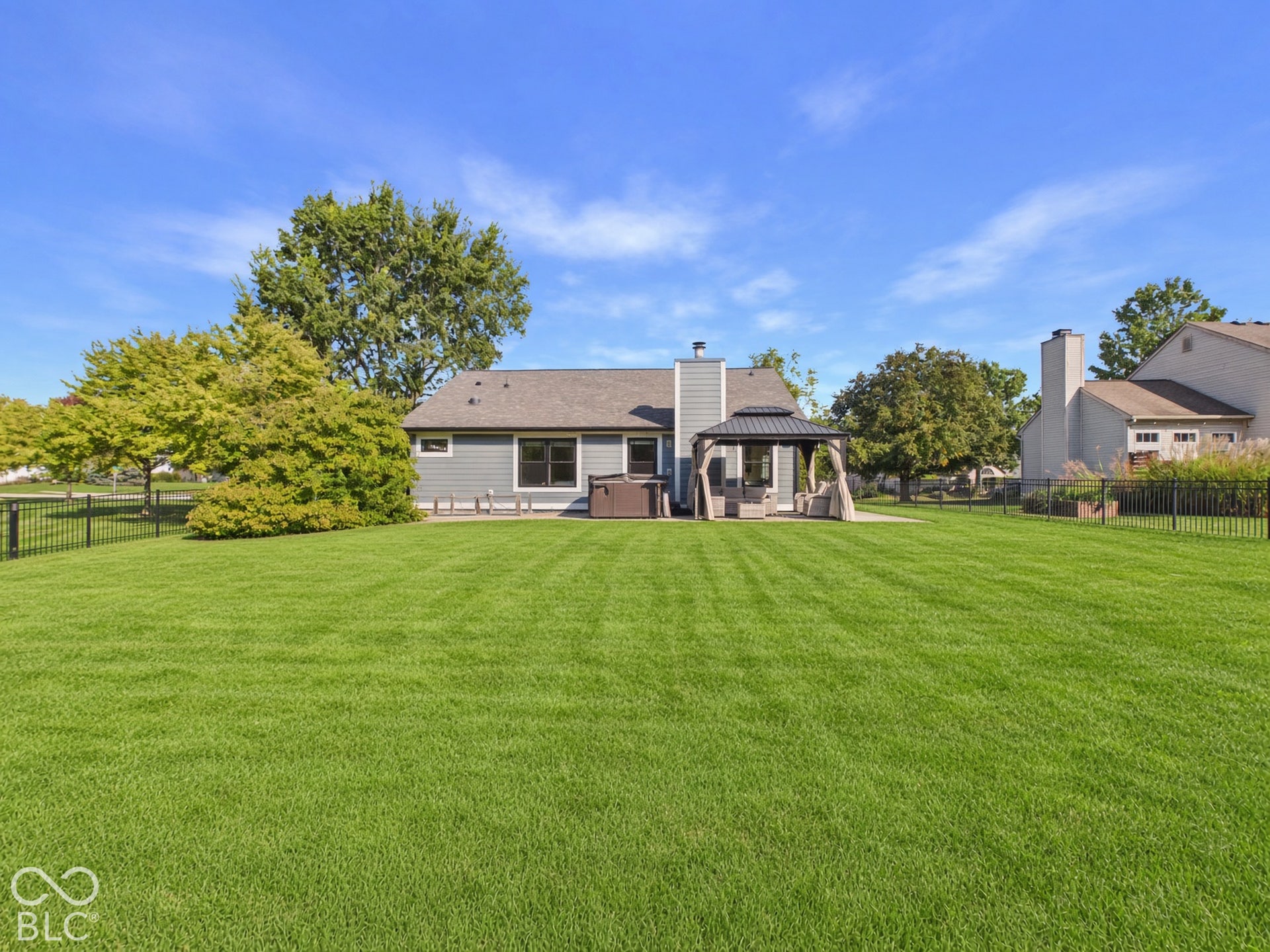 Expansive green lawn with a black metal fence, leading to a home featuring grey siding, a dark roof, and a covered patio structure