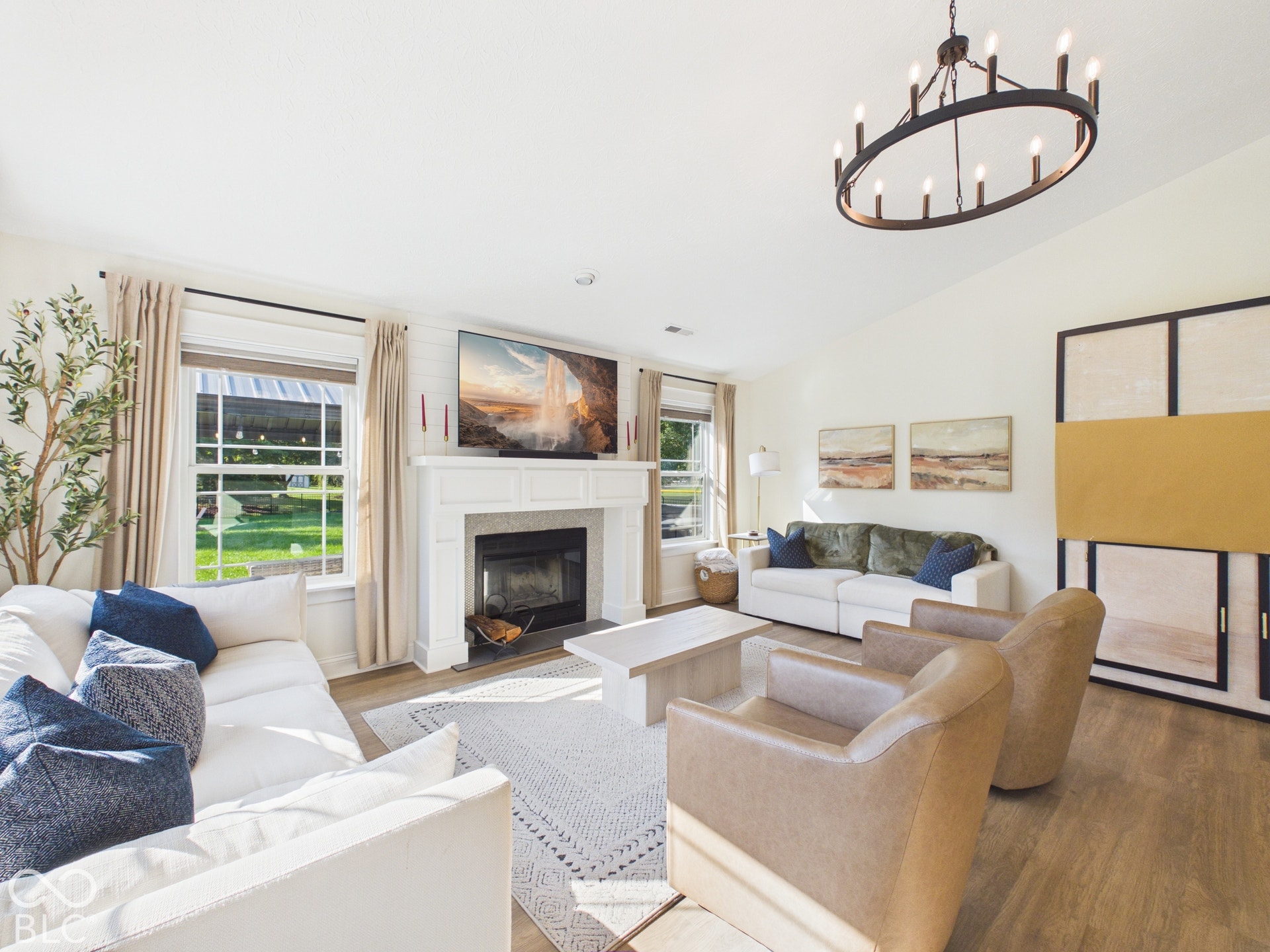 Vaulted ceiling with an oval chandelier, wood-finish flooring, white shiplap accent wall, fireplace with white mantel, and large windows