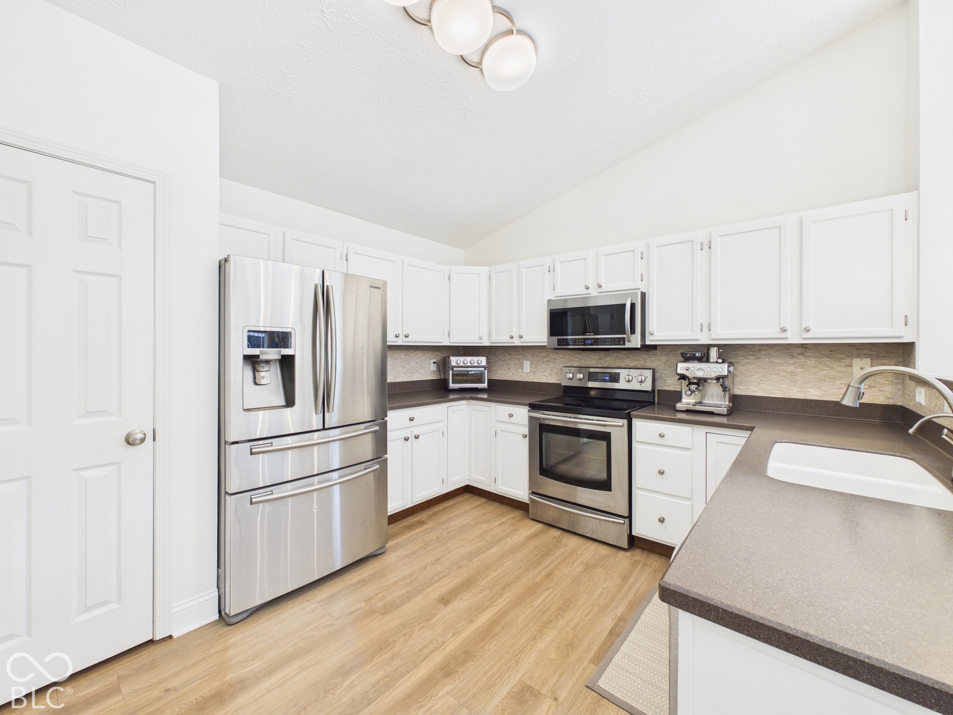 Kitchen featuring vaulted ceiling, wood-finish flooring, white cabinetry, stainless steel appliances, and a composite sink