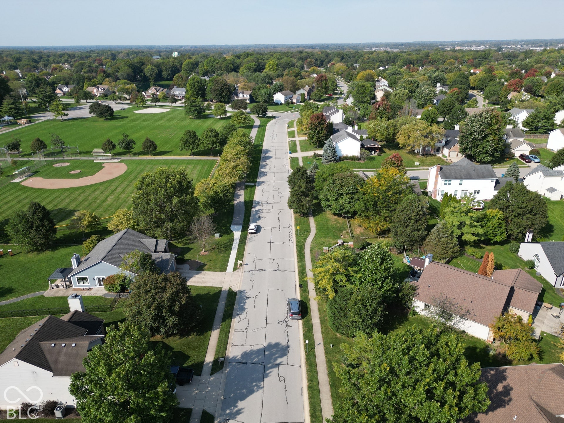 Established residential area with mature trees, paved streets, and a manicured park with a baseball diamond