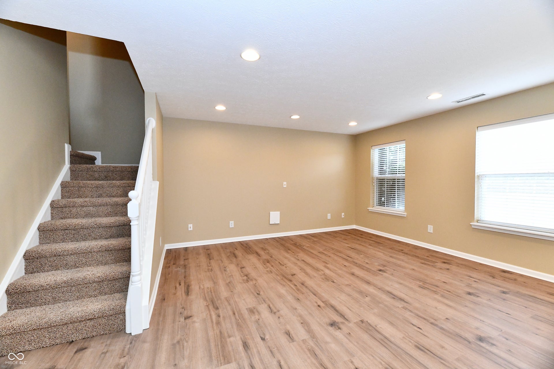 Recessed lighting, wood-finish flooring, two windows with blinds, and a carpeted staircase with white railings