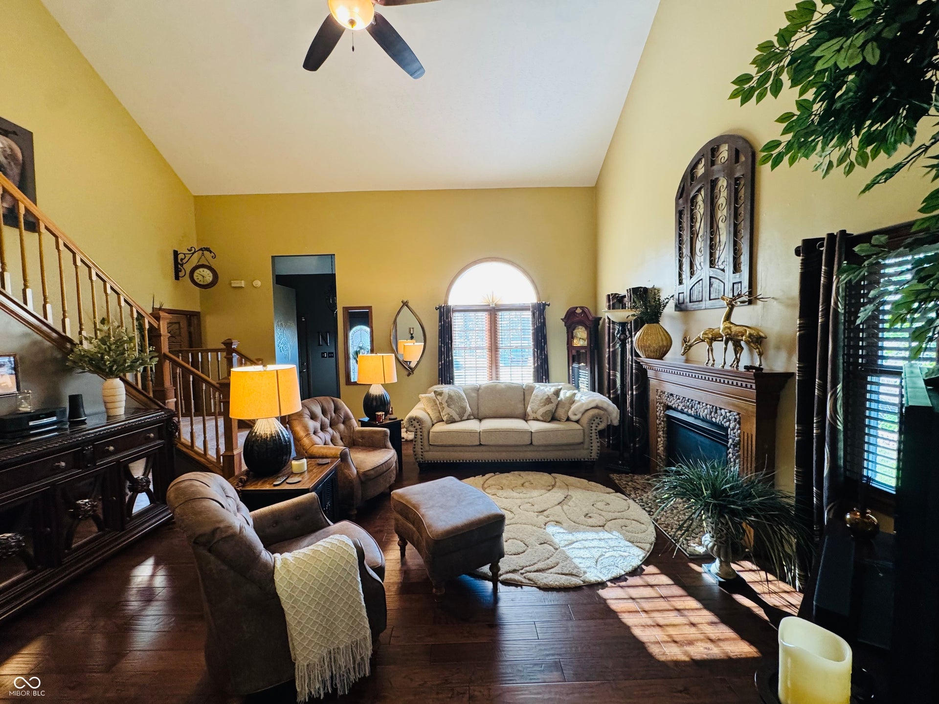 Vaulted ceiling with a ceiling fan, wood-finish flooring, staircase with wood railing, arched window, and a fireplace with an ornate wood mantel