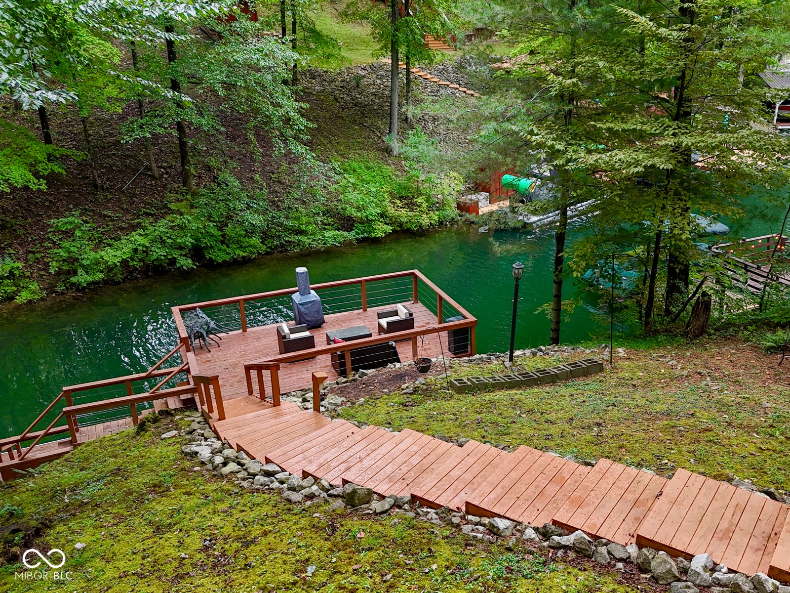 Multi-level wood deck with cable railing, wood steps to the water, and mature trees surrounding the property