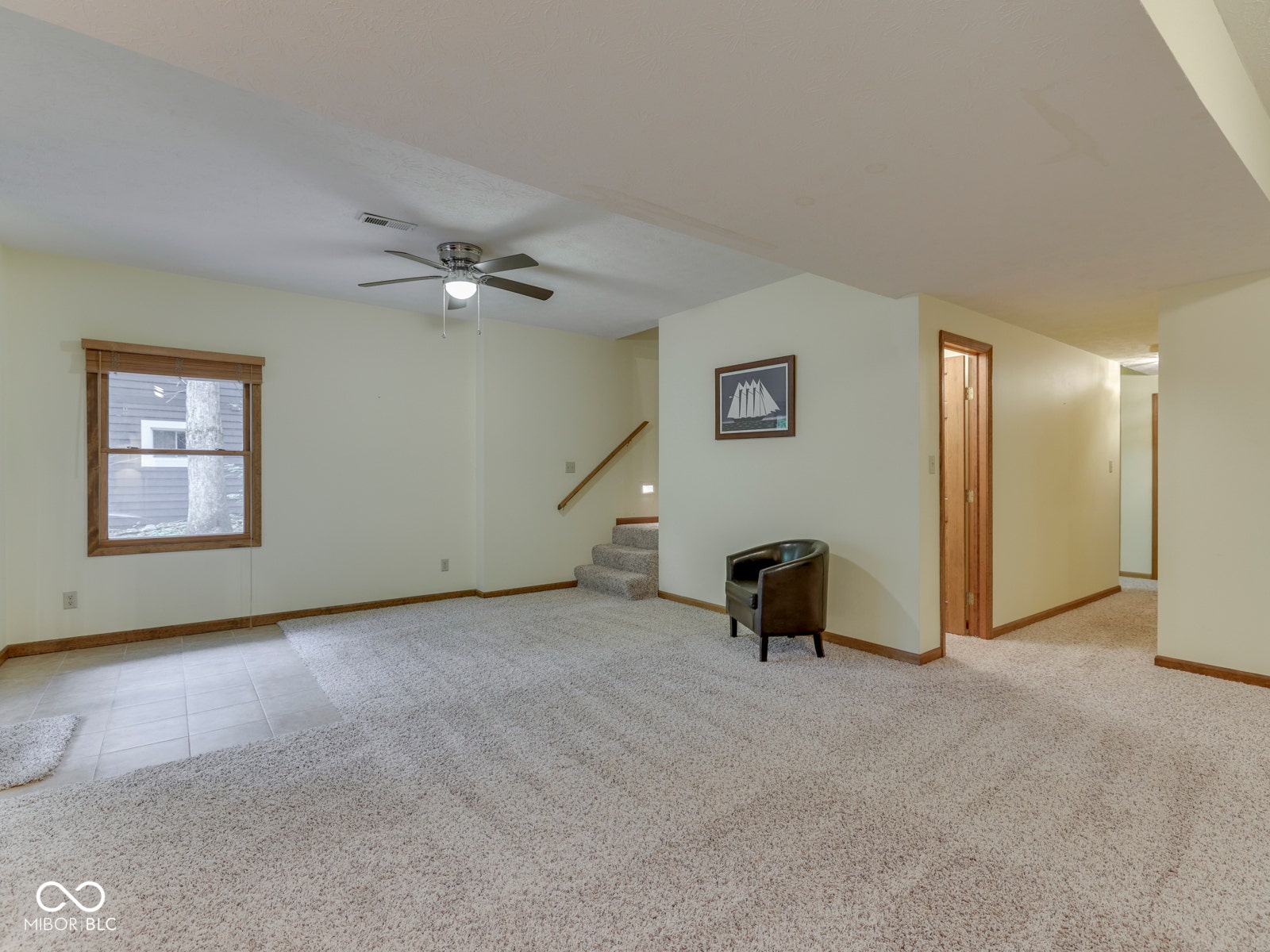 Spacious living area featuring neutral carpeting, a ceiling fan, a window with wood trim, and a staircase with a wood handrail