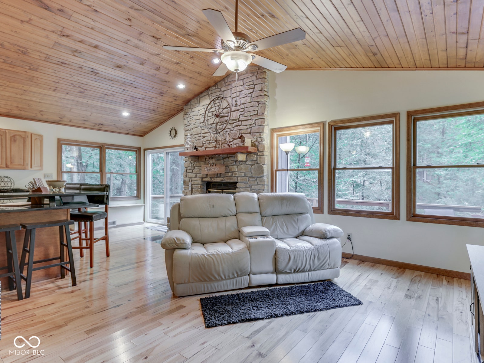 Vaulted wood-paneled ceiling with a ceiling fan, stone-clad fireplace with a wood mantle, wood-finish flooring, and multiple windows with wood trim