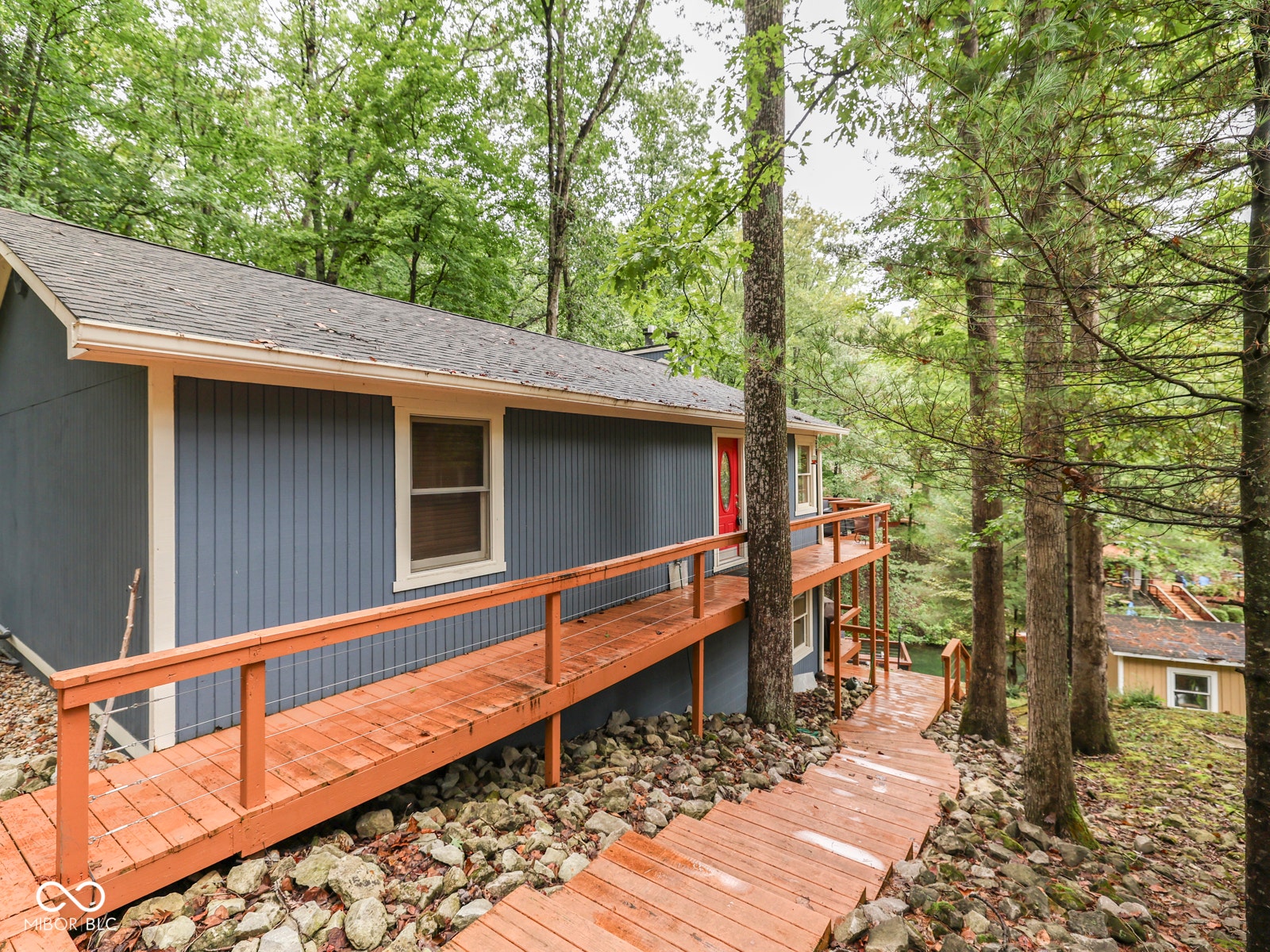 Blue vertical siding exterior with a red entry door, a shingled roof, an elevated wood deck, and a wood ramp