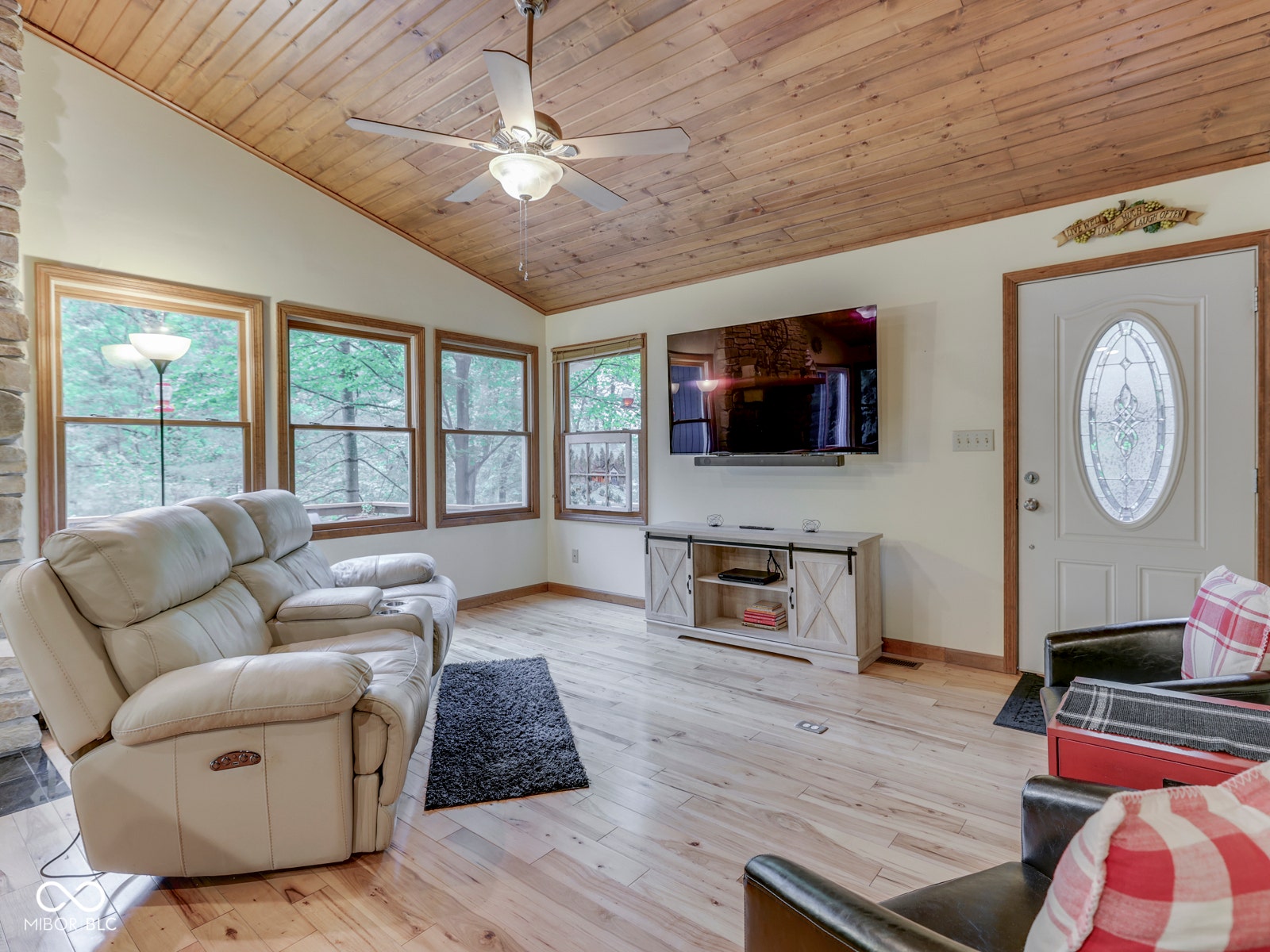 Vaulted wood-paneled ceiling with ceiling fan, light wood-finish flooring, white paneled door with oval leaded glass insert, multiple windows with wood trim, and a stone-clad fireplace surround