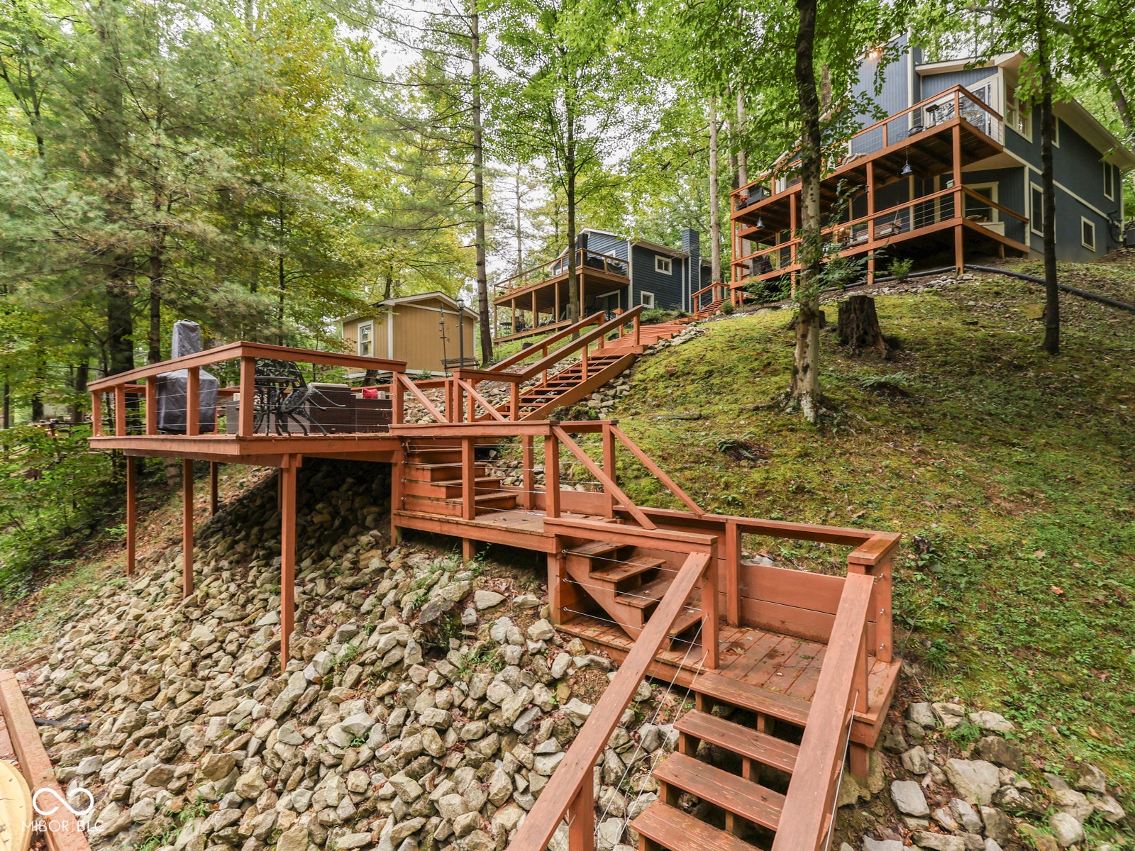 Multi-level decks with wood railings, exterior stairs with wood treads, and a sloped terrain with natural rock landscaping