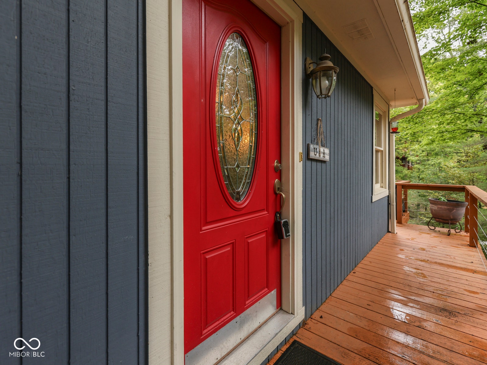 Red entry door with an oval glass insert, vertical panel siding, exterior wall sconce, and a wood-finish deck with cable railing