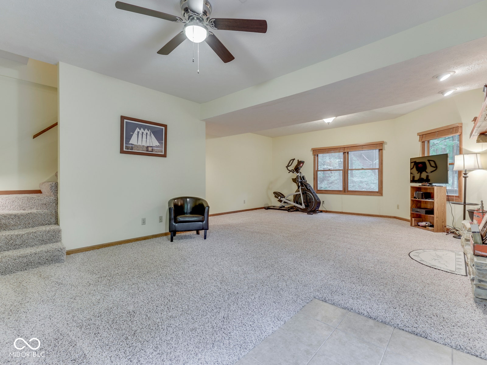 Spacious room featuring light-toned carpeting, a ceiling fan with integrated lighting, and multiple windows with wood trim