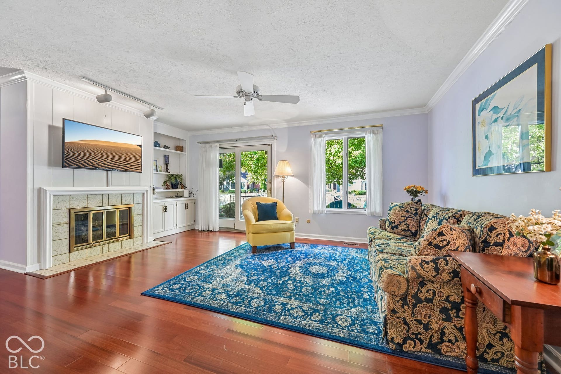 Wood-finish flooring, built-in shelving, stone-surround fireplace, large windows, and a ceiling fan