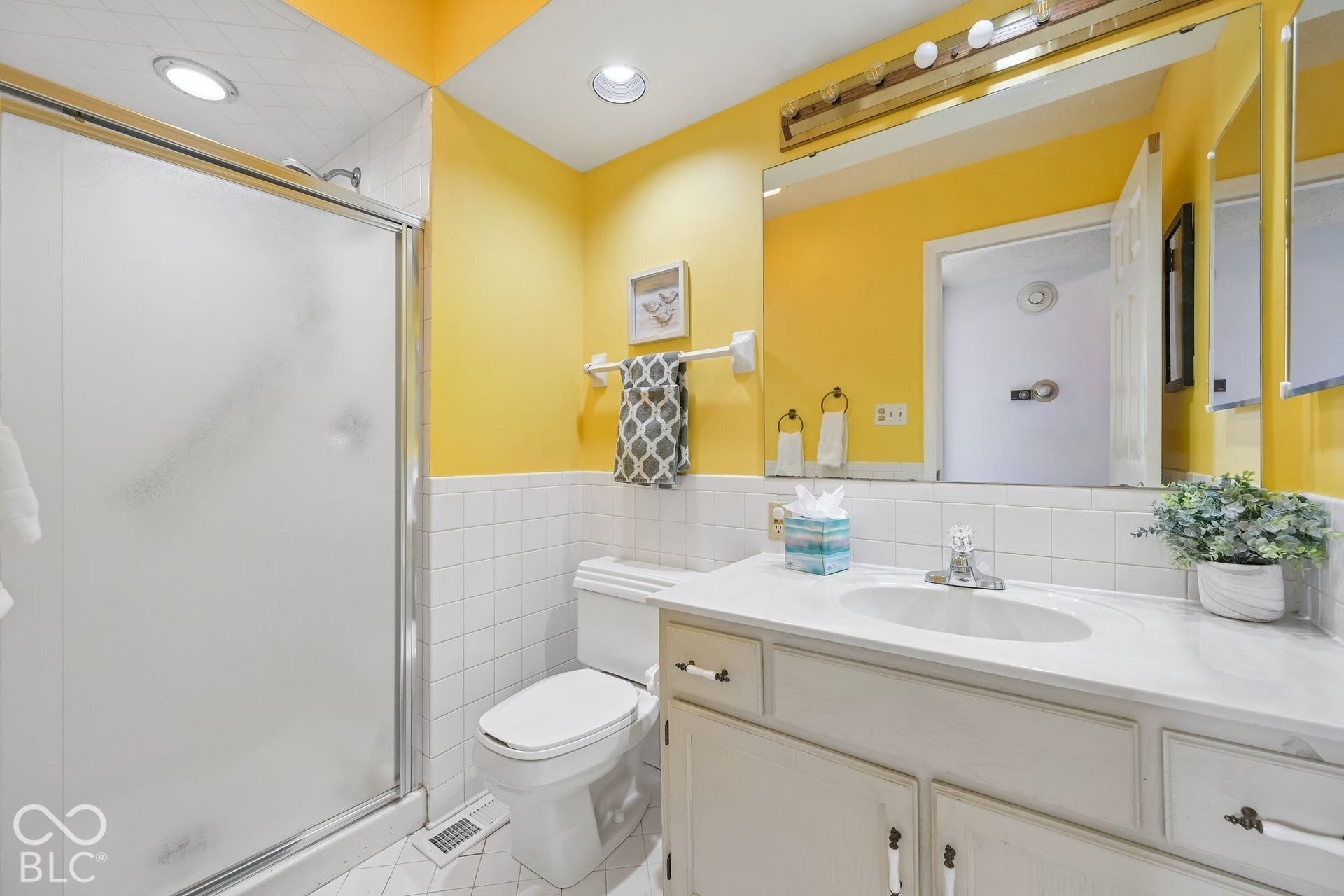 White tile-surround shower with sliding glass door, single vanity with integrated sink, chrome faucet, and expansive wall-mounted mirror