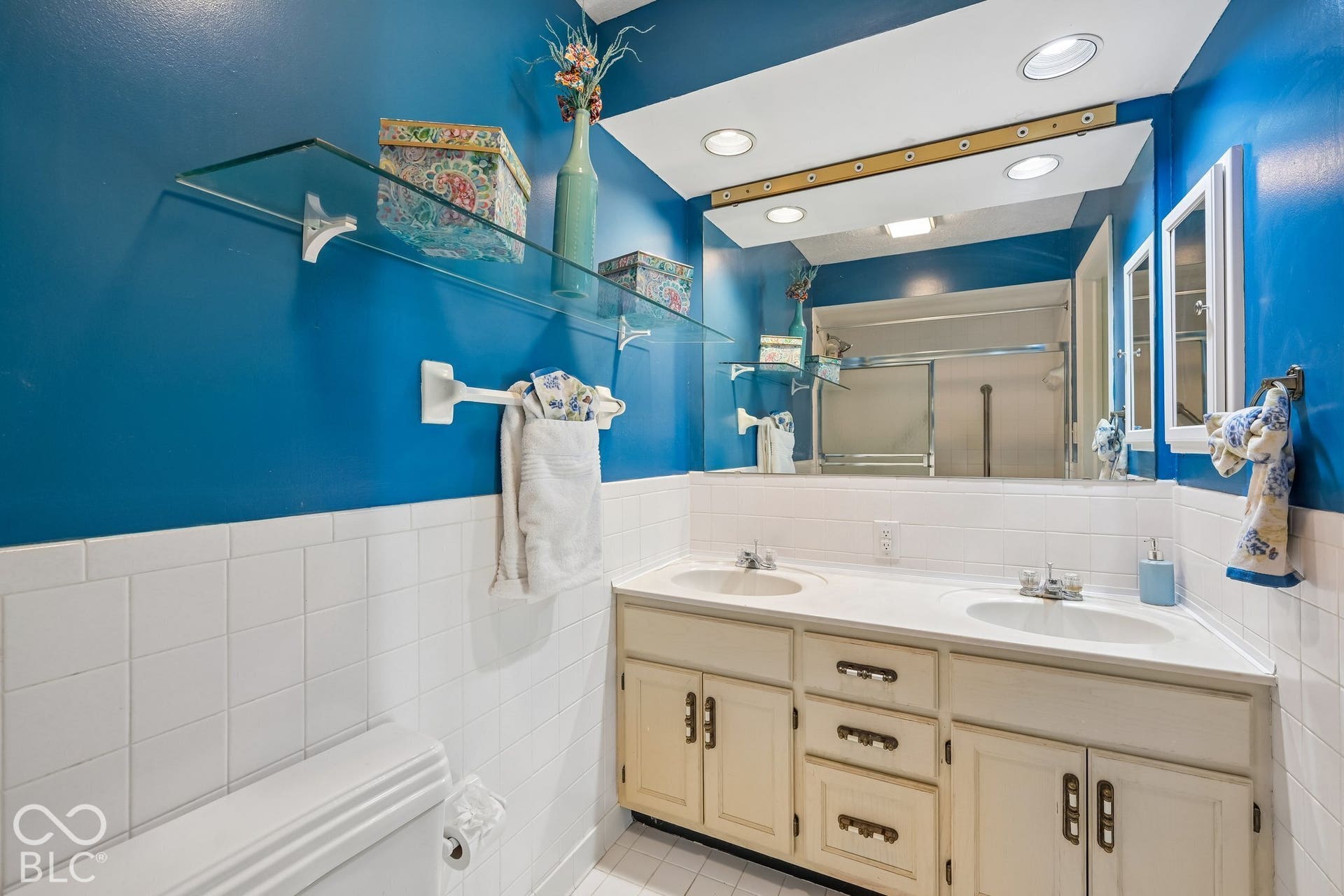 Double vanity with integrated sinks, ample storage cabinetry, glass shelving, and recessed lighting