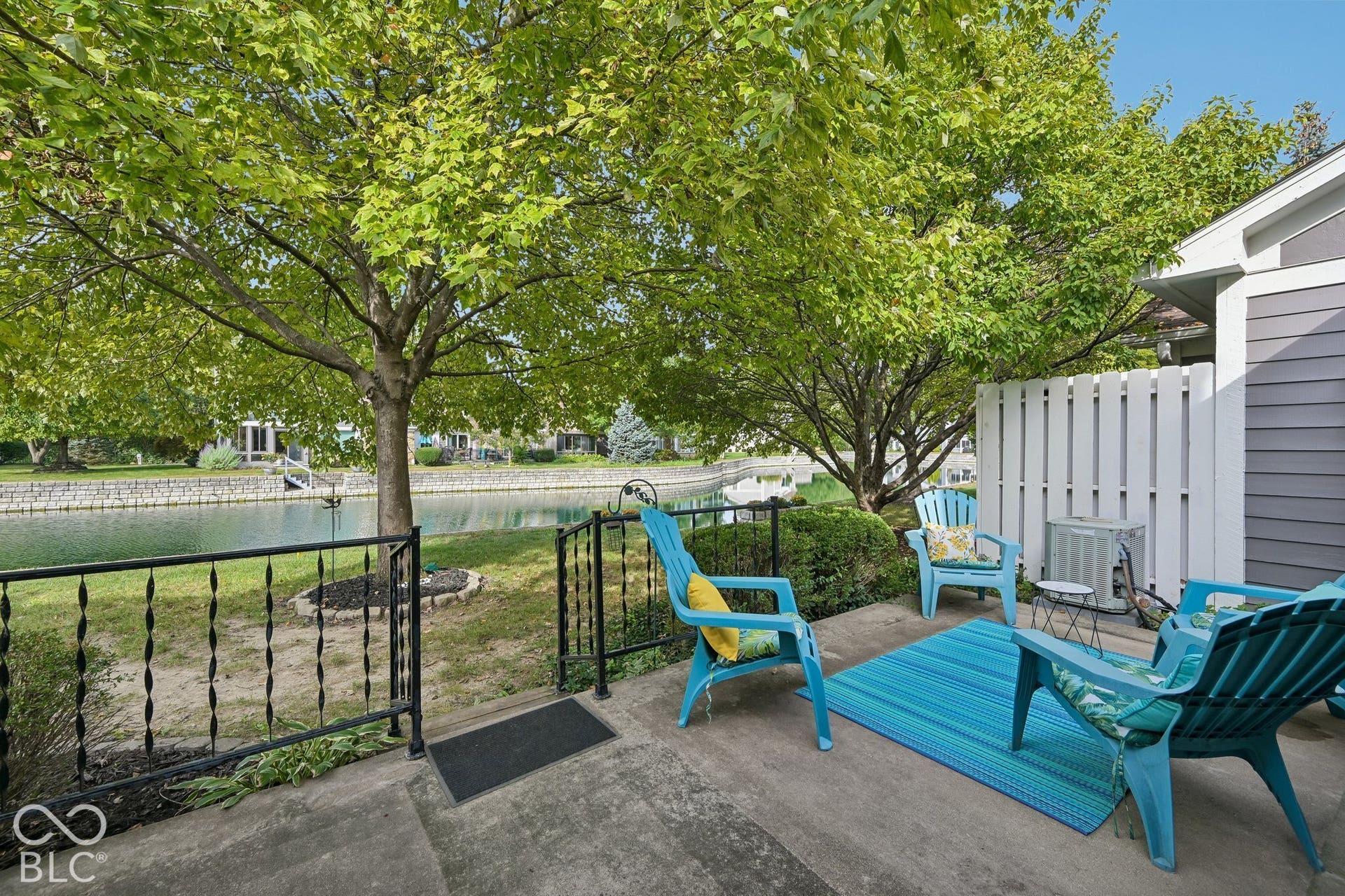 Concrete patio area with a white privacy fence, mature trees, and a black wrought iron railing, overlooking a water feature