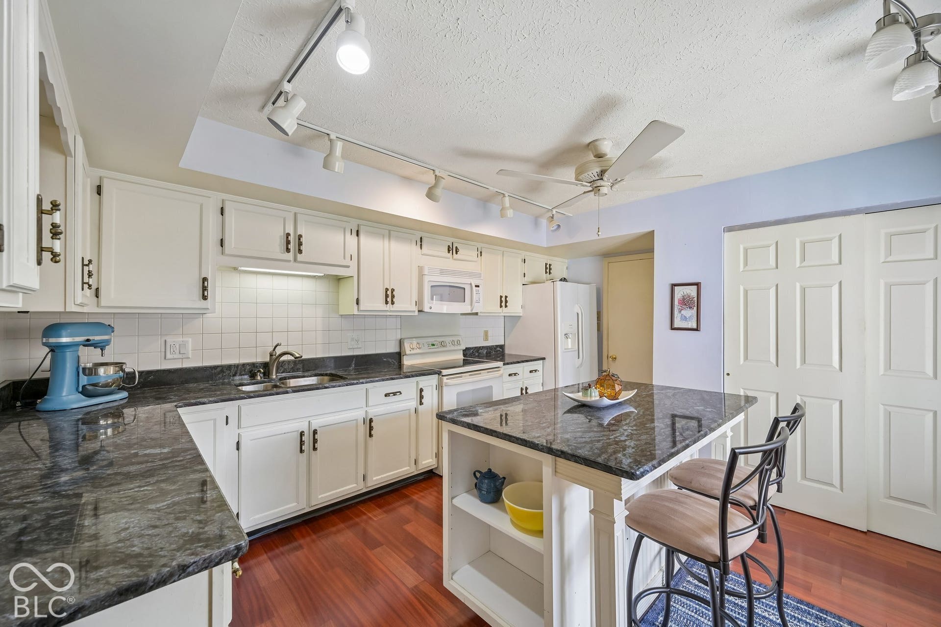 White cabinetry, dark stone countertops, wood-finish flooring, central island with seating, and white appliances
