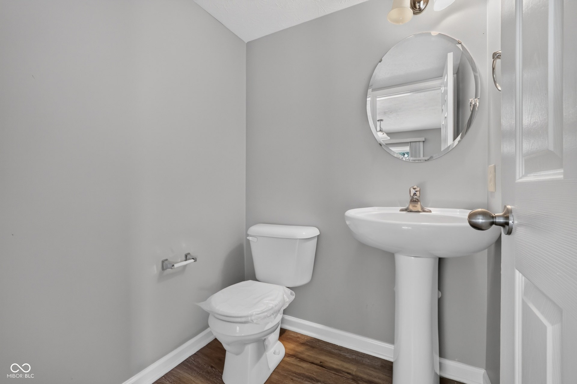 Pedestal sink with a circular mirror, brushed nickel faucet, and wood-finish flooring