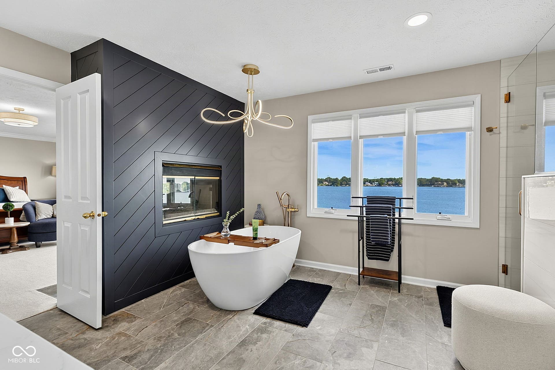 Primary ensuite with freestanding soaking tub with a modern chandelier, a built-in fireplace feature wall, a glass-enclosed shower, and large windows with water scenery.